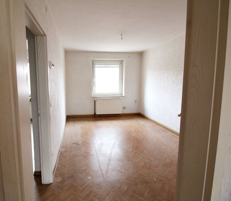 Leeres, unmöbliertes Zimmer mit Holzboden, weißen Wänden und einem einzigen Fenster.