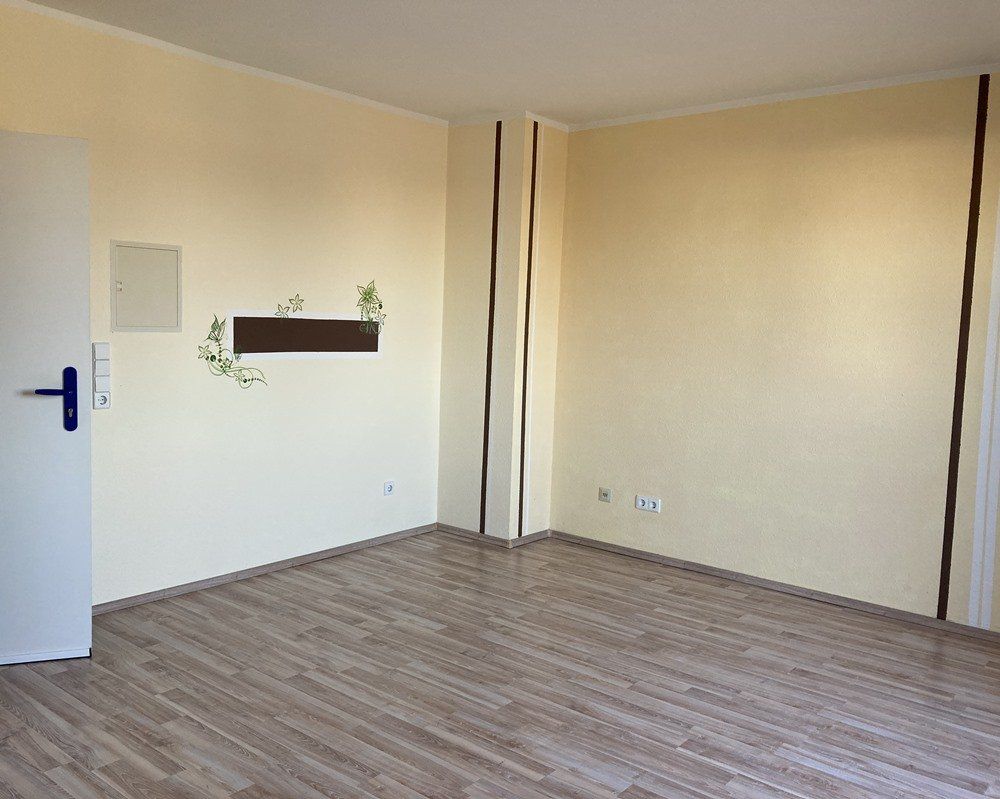 Leeres beigefarbenes Zimmer mit Holzbodenoptik, offener Tür und kleinem Wandregal mit Pflanzen.