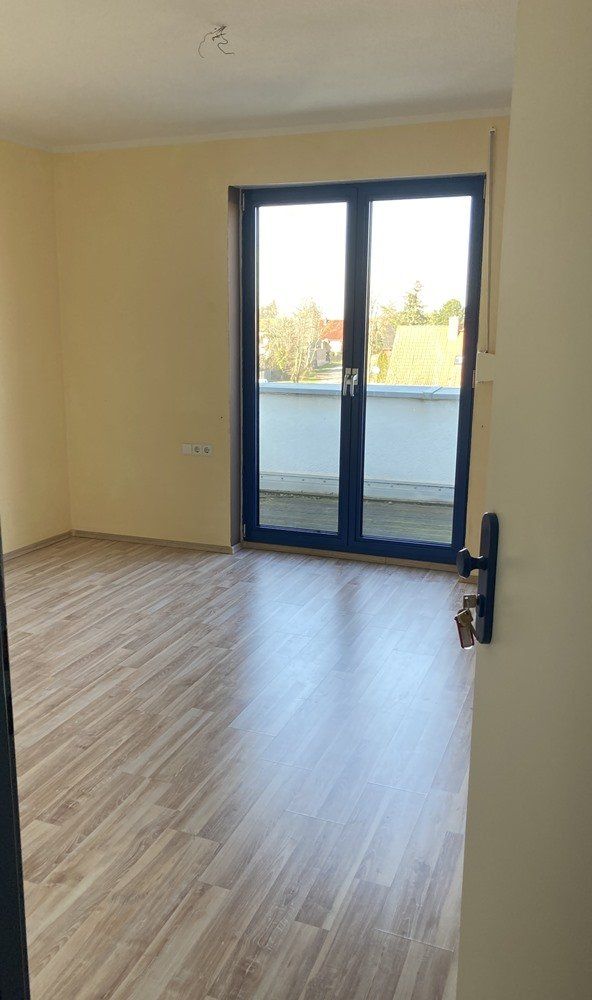 Leeres beigefarbenes Zimmer mit Holzbodenoptik und Glasbalkontüren, die Tageslicht hereinlassen