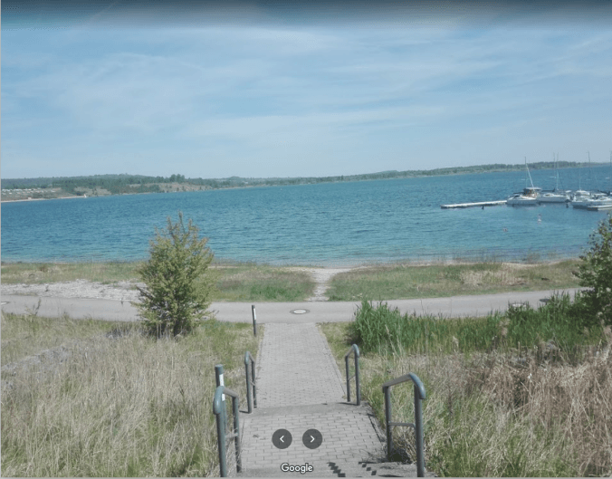 Blick die Stufen hinunter auf einen Sandstrand und blaues Wasser mit Booten und einer fernen Küstenlinie unter klarem Himmel