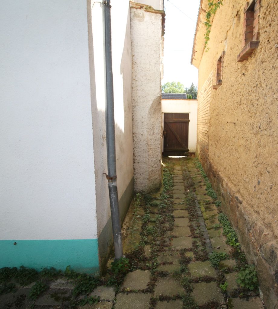 Eine schmale, mit Kopfsteinpflaster belegte Gasse zwischen weißen und beigen Mauern, die an einem Holztor endet.