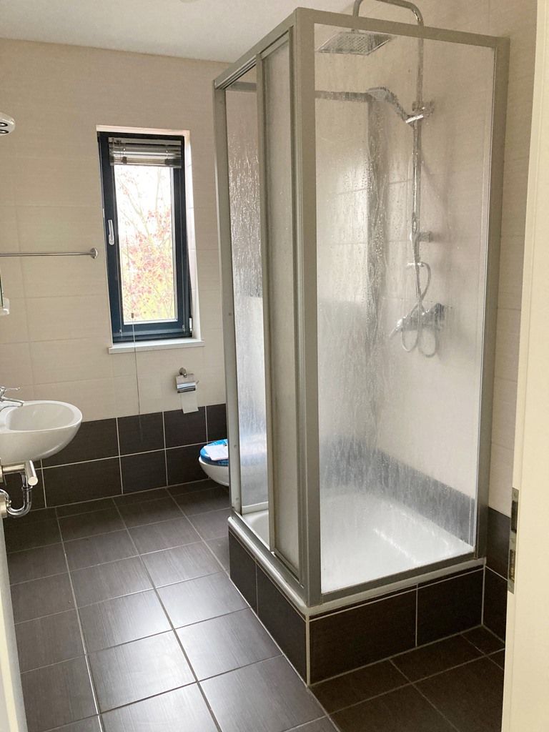 Barrierefreies Badezimmer mit ebenerdiger Dusche, Milchglastür, weißem Waschbecken und dunklem Fliesenboden.