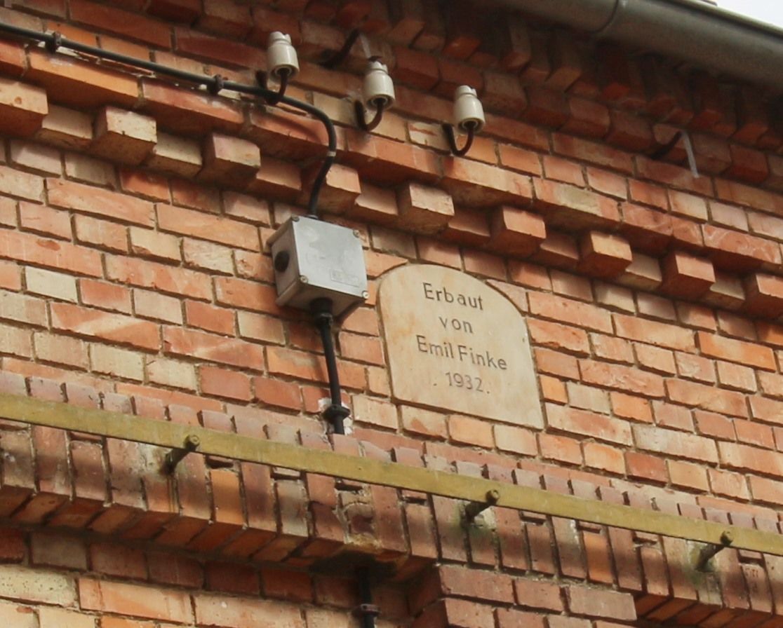 Alte Backsteinmauer mit einem kleinen Verteilerkasten, Rohren und einem verwitterten Schild unter einer Dachkante