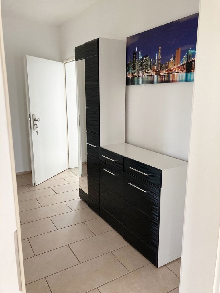 Moderner Flur mit weißen Wänden, Fliesenboden, schwarz-weißen Schränken, offener Tür und Wandbild mit Stadtsilhouette.