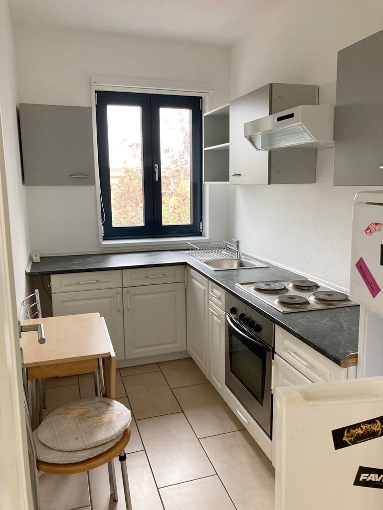 Kleine weiße Küche mit grauen Schränken, schwarzem Fenster, Herd, Spüle sowie einem Holztisch und Hocker.