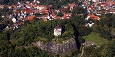 Luftaufnahme einer Burg auf einem Hügel über einer bewaldeten Klippe und einem Dorf mit roten Dächern