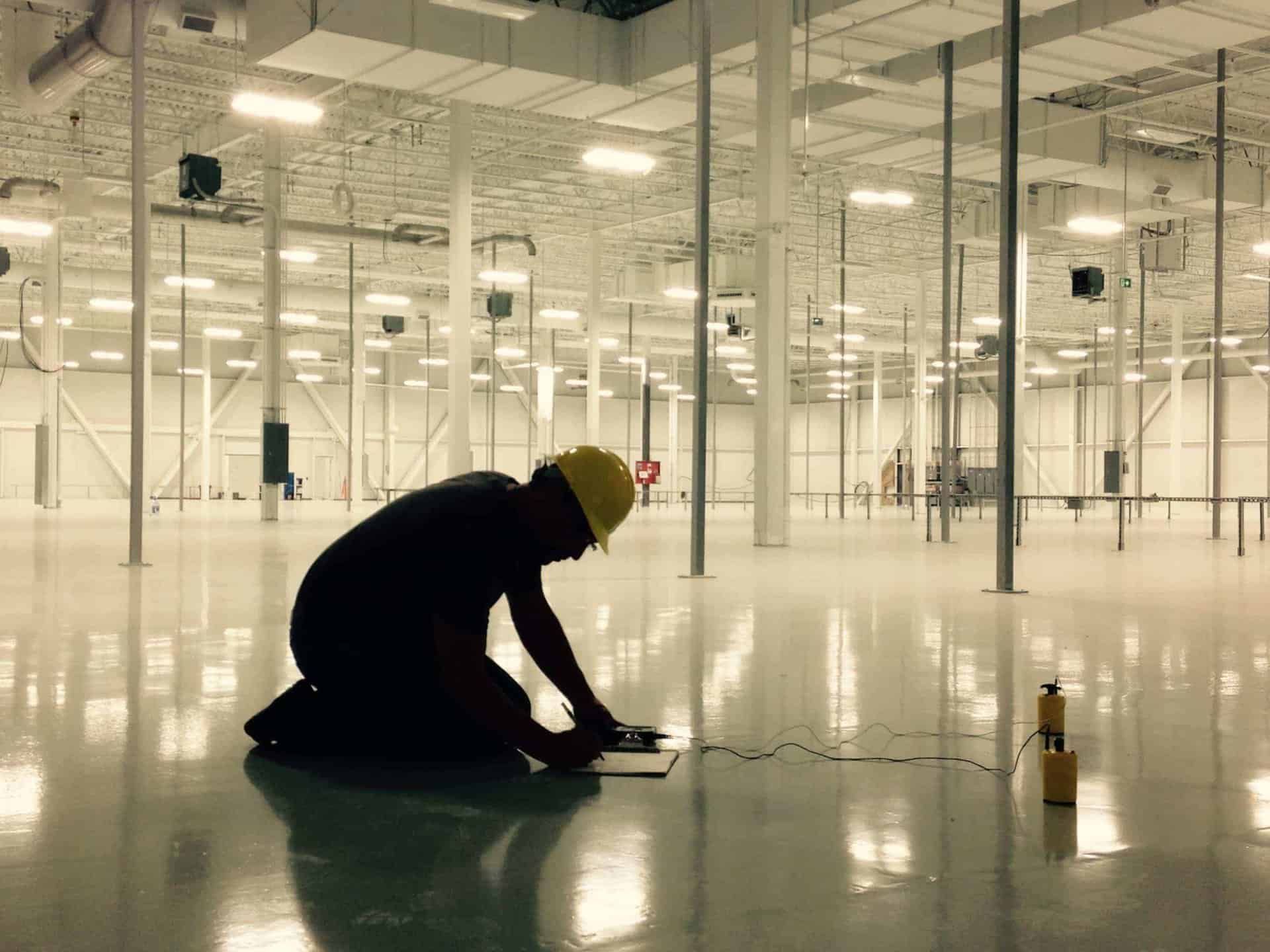 Worker kneels in a bright empty warehouse, inspecting a device on the floor.