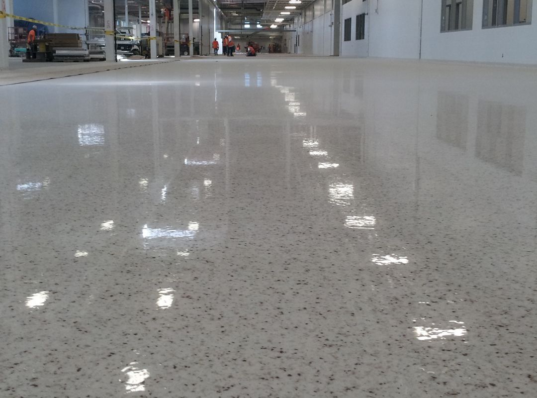 Polished speckled epoxy floor in a bright industrial warehouse aisle with overhead lights reflecting on the surface