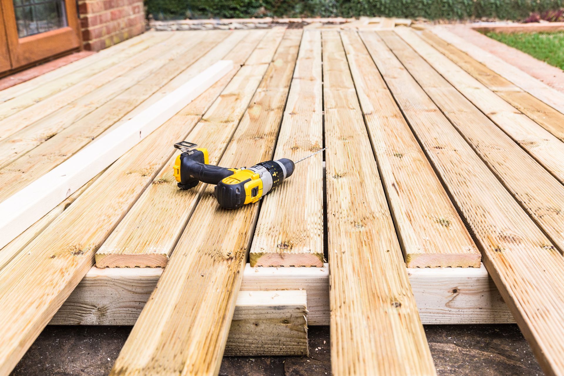 A yellow and black power drill sits on partially installed wooden decking boards outdoors near a house.