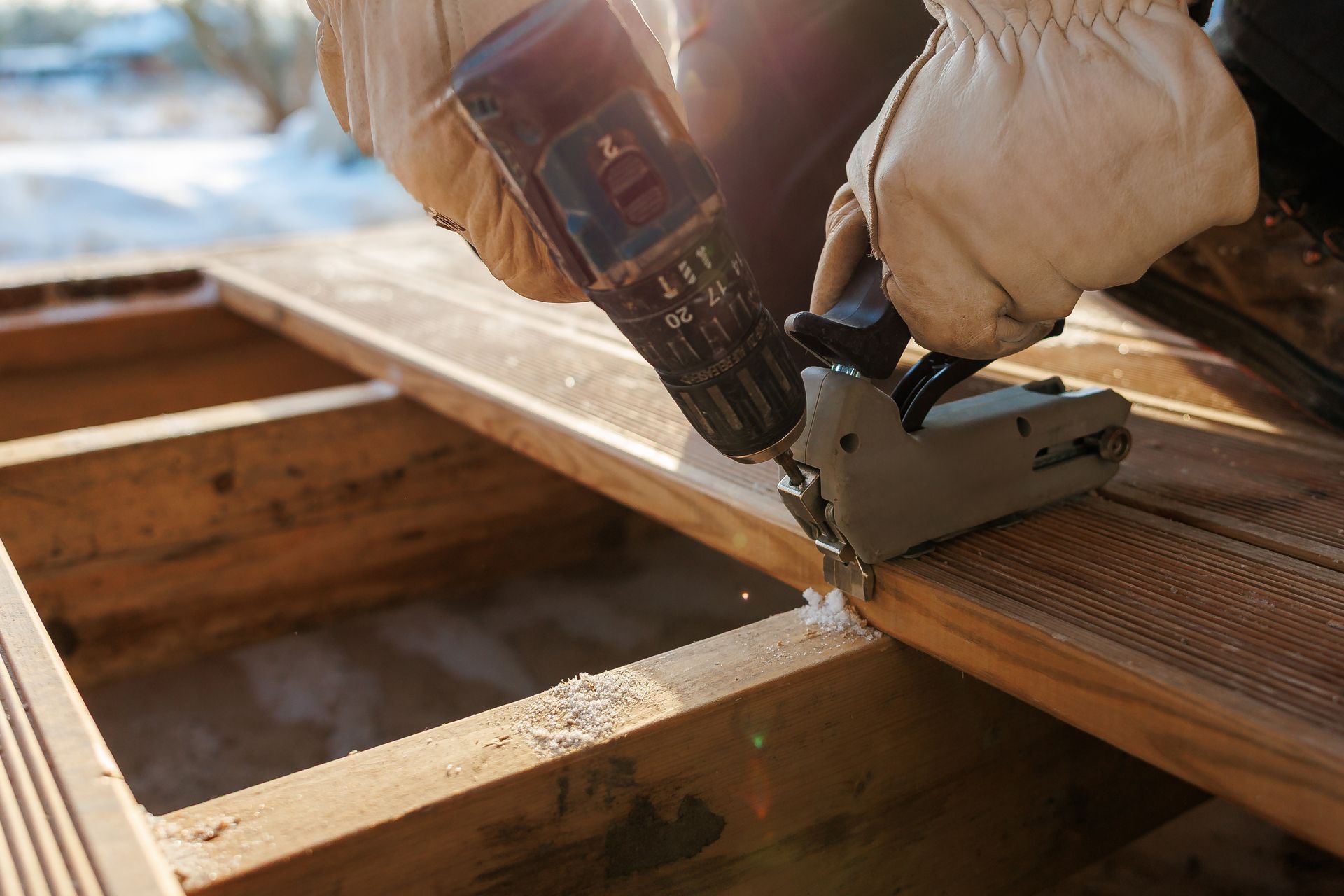 A person wearing work gloves uses a drill attachment to secure wooden deck planks to a frame.