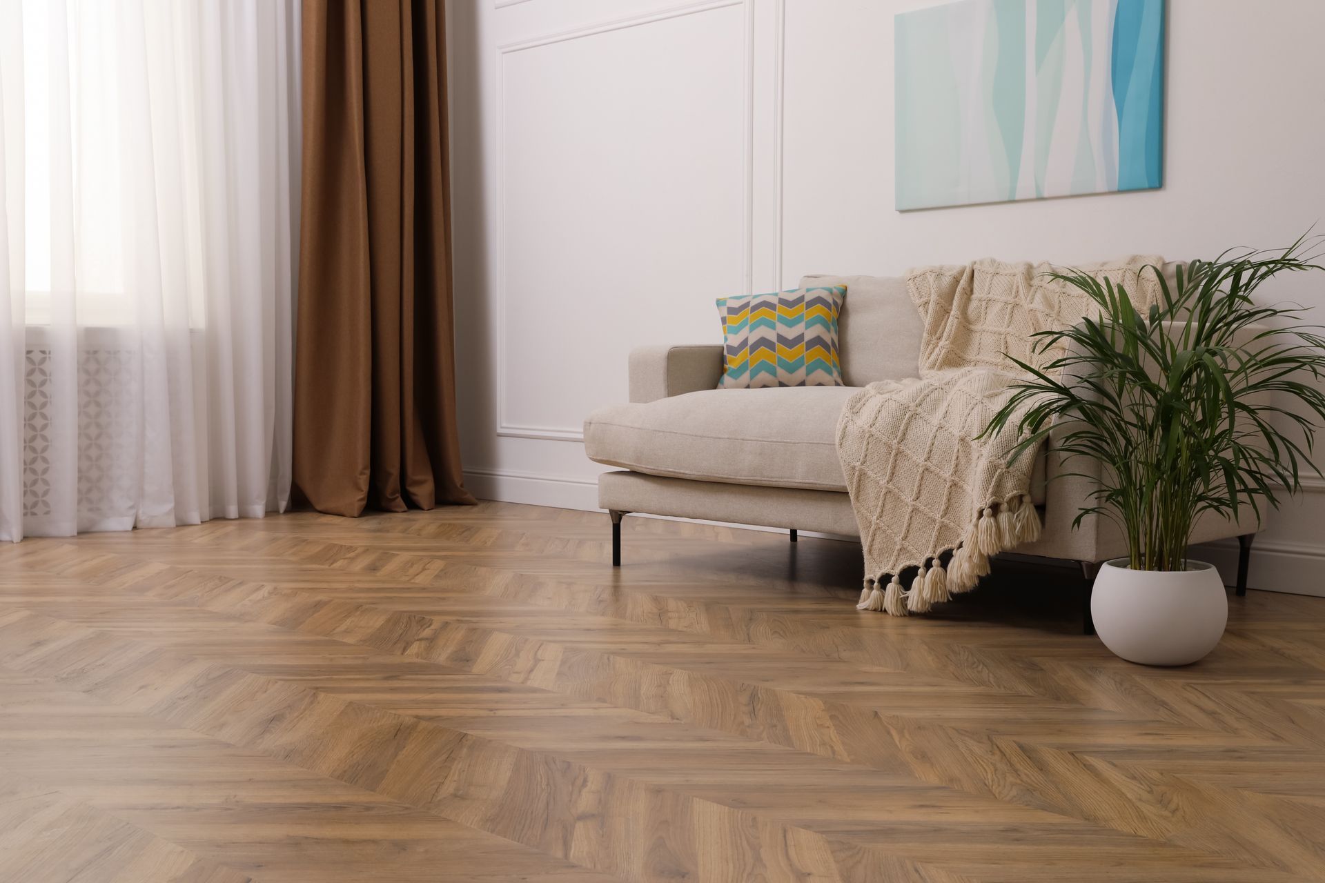 A beige sofa with a throw blanket and patterned pillow sits on herringbone wood flooring next to a potted plant.