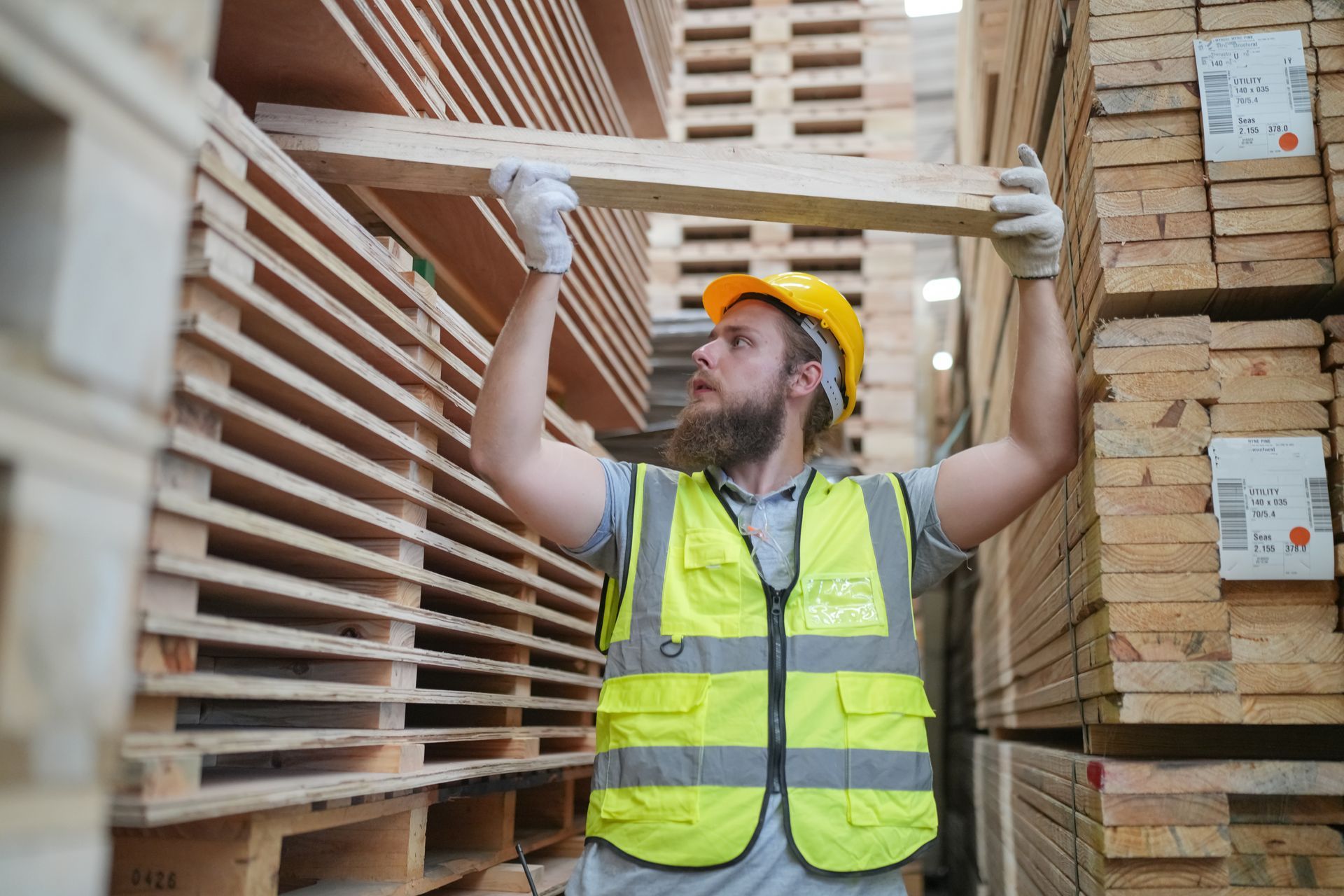 A warehouse worker in a yellow hard hat and high-visibility vest lifts a wooden board while working near stacked lumber.