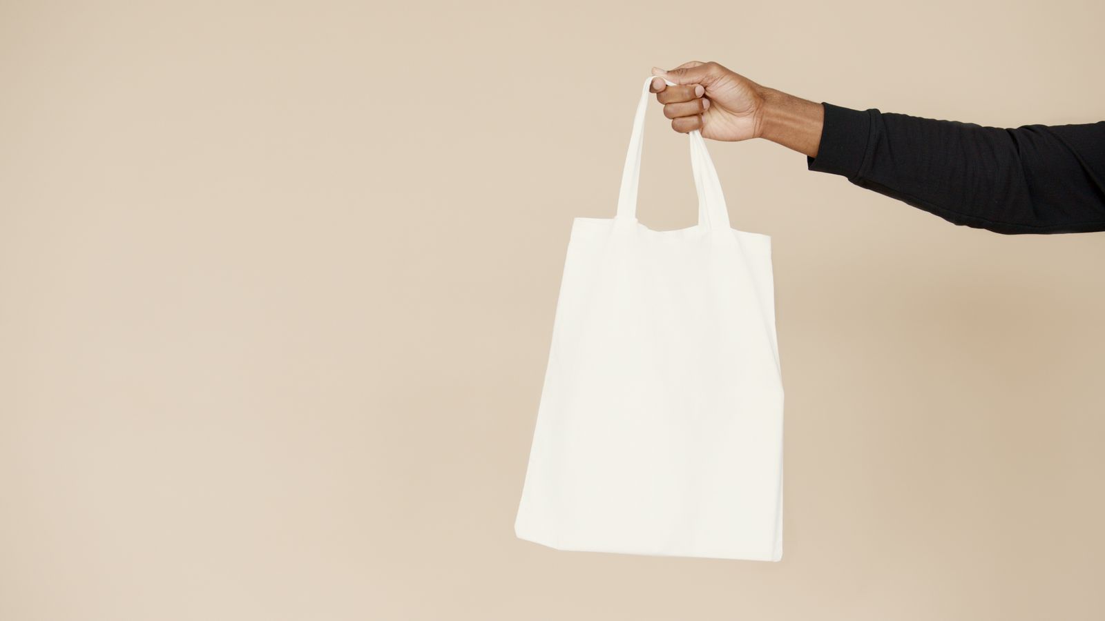 Hand holding a plain white tote bag against a beige background