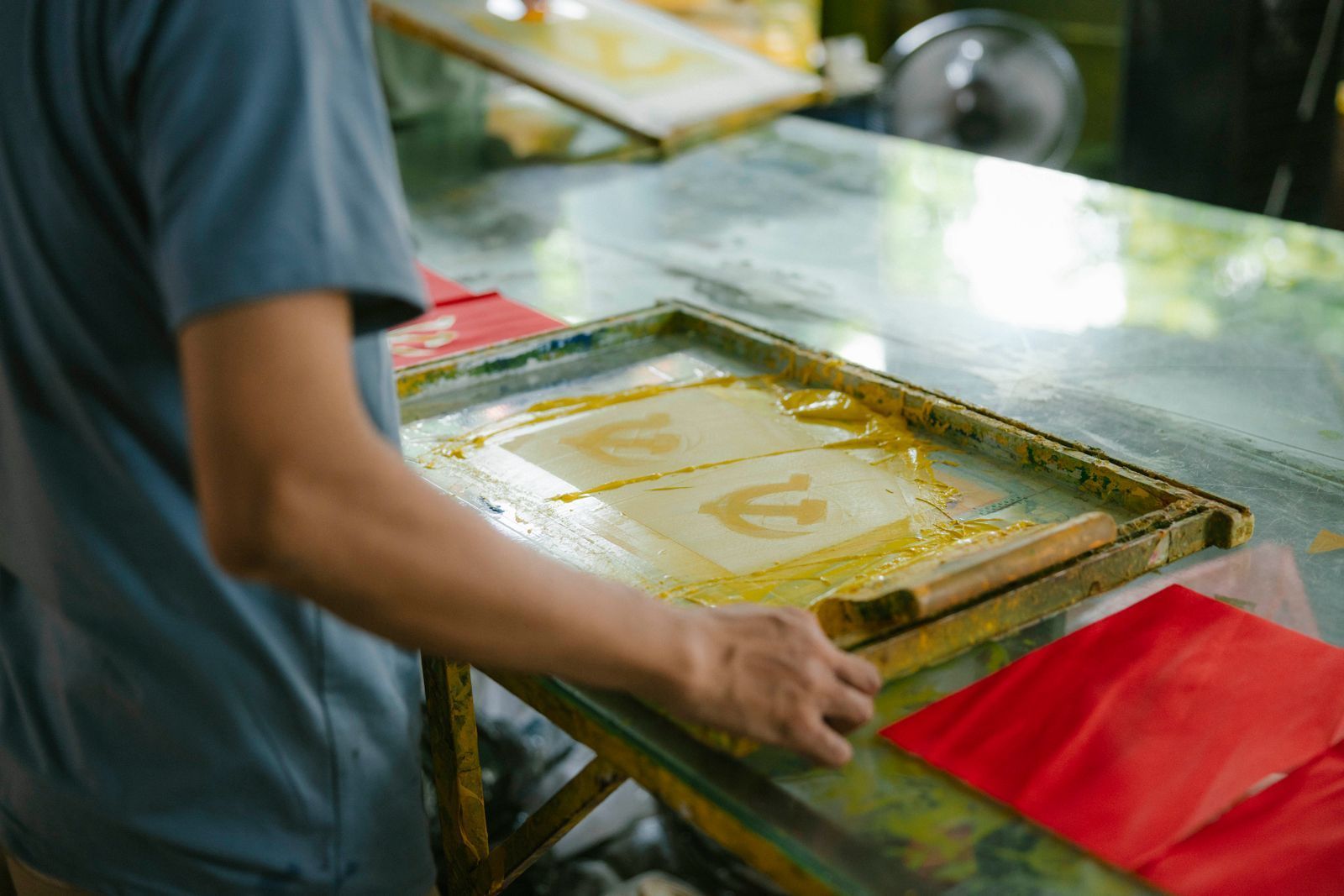 Person screens a yellow design on a worktable with red cloths in a workshop