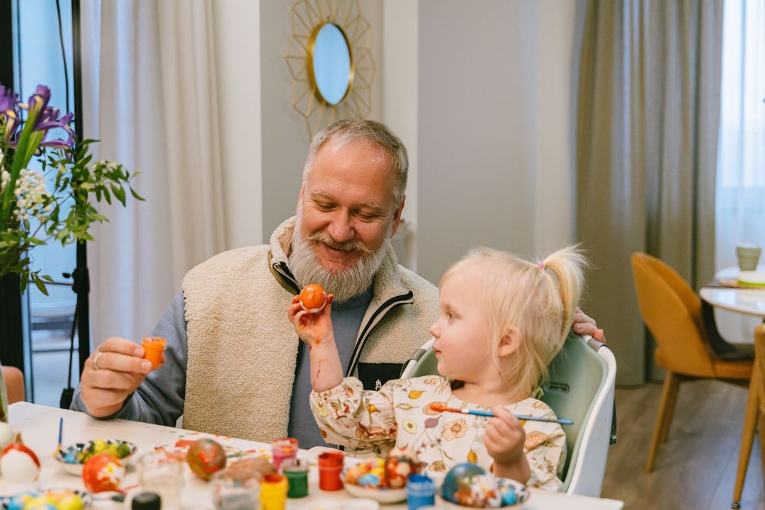 Man and child smiling at a table, painting eggs with colorful supplies in a bright dining room