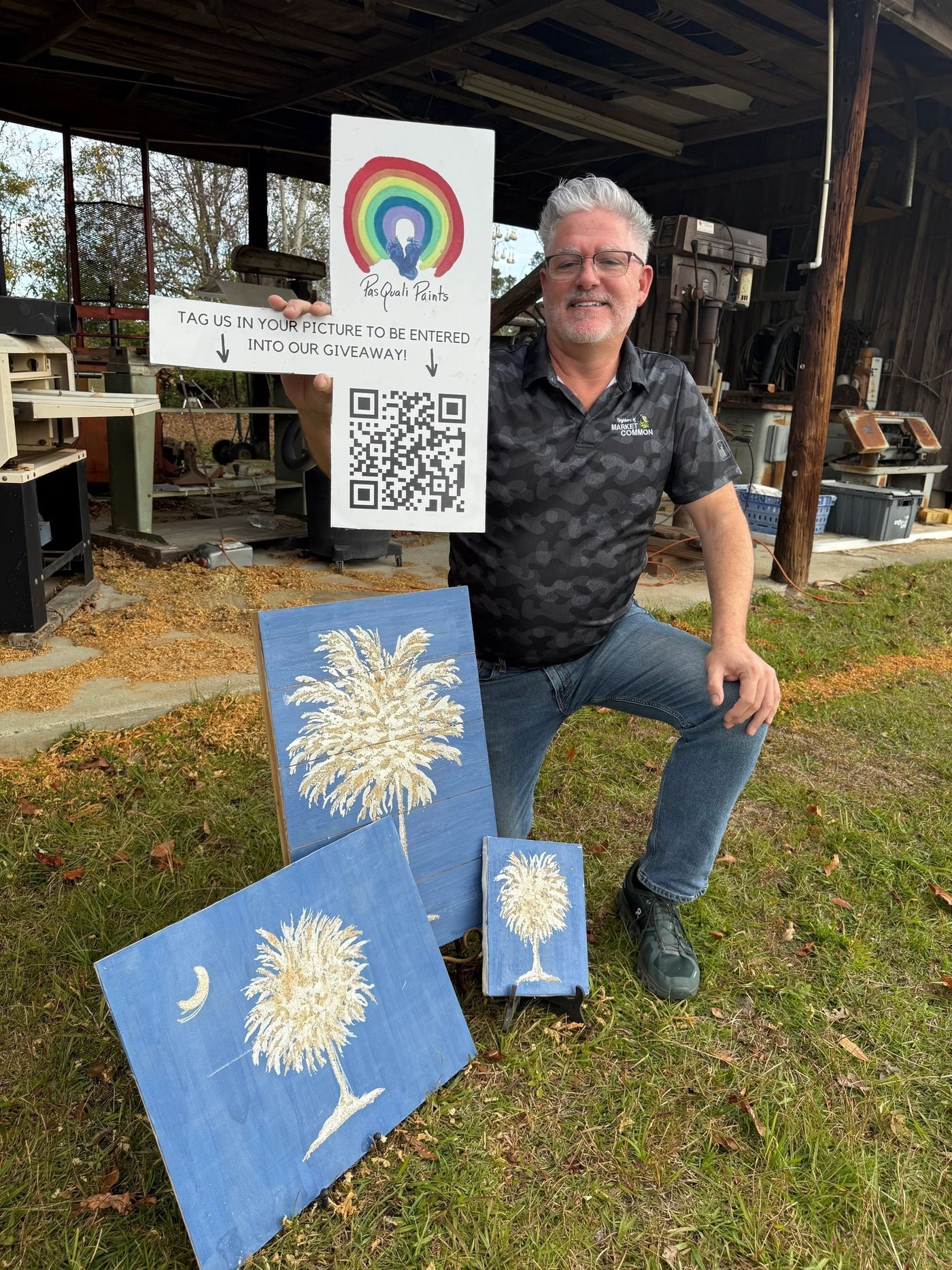 Man posing beside colorful artwork and a QR code under a rustic shelter.