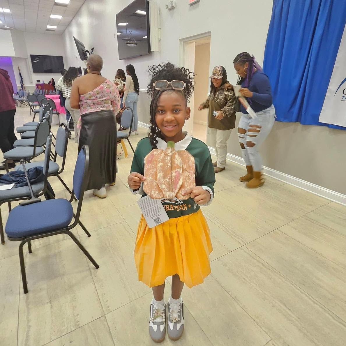 Smiling child holding a paper fan at an indoor event, wearing a yellow skirt and green sweater.