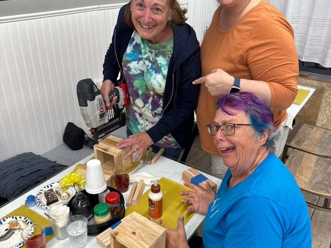 Three people smiling at a table with art supplies and craft items at a community event