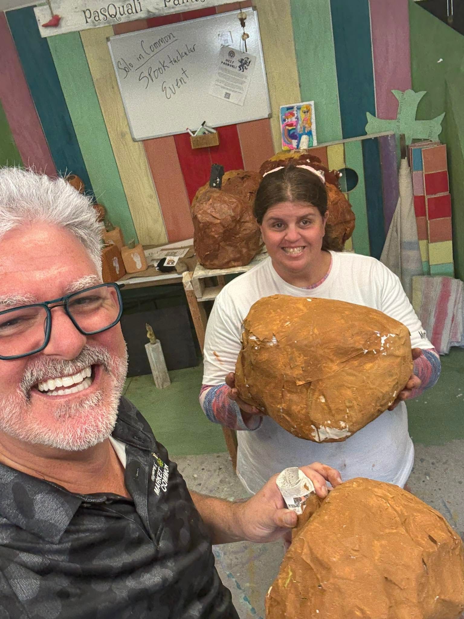 Two people smiling in a colorful studio, holding large tan clay pieces.