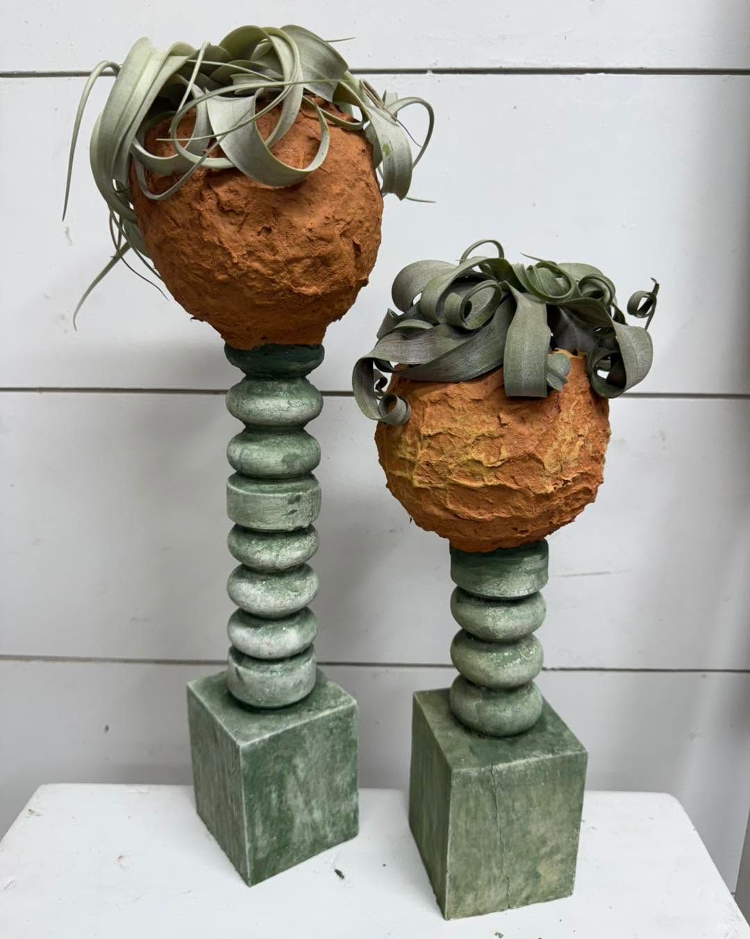 Two decorative plant sculptures in green and brown on square bases against a white wall