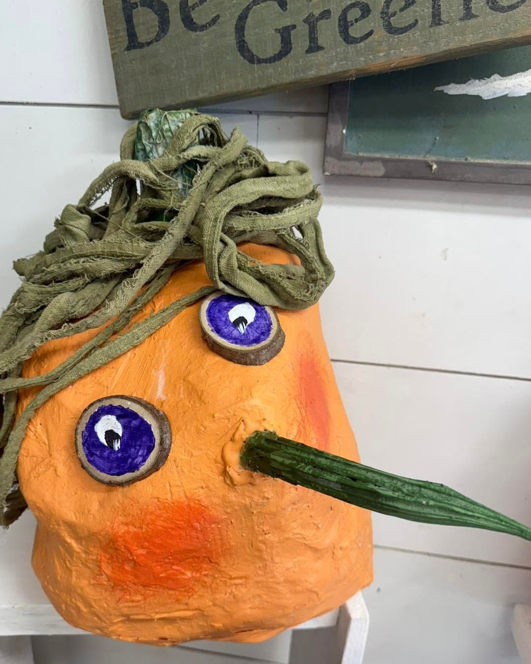 Orange handmade face with blue eyes, green yarn hair, and a long green nose on a wall display