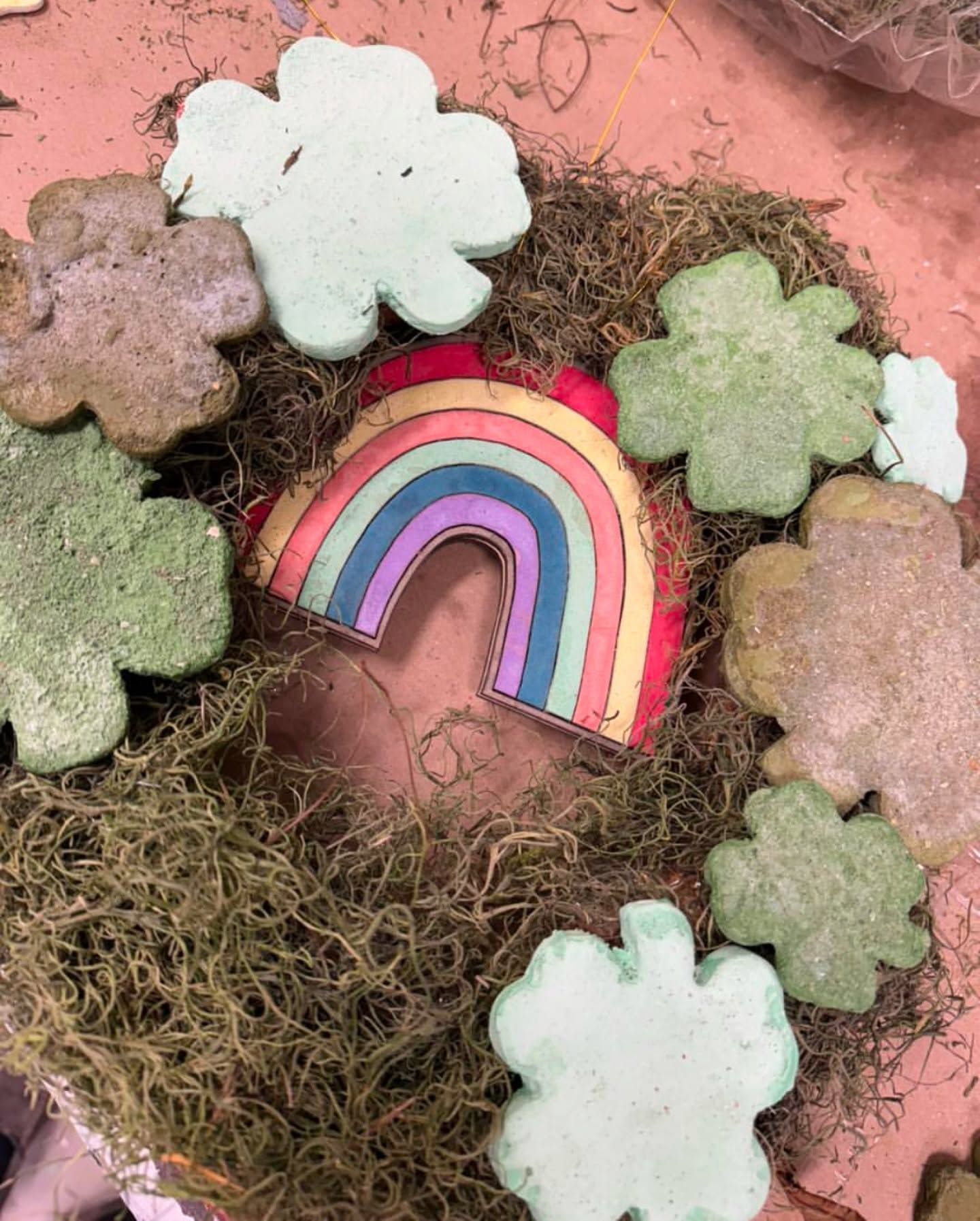 Rainbow ornament surrounded by pastel flower shapes on a pink, mossy background