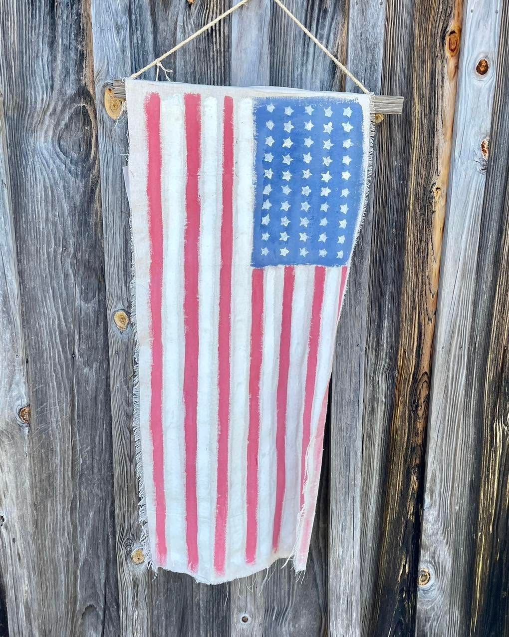 Hand-painted American flag on a small sign hanging against weathered wooden boards.