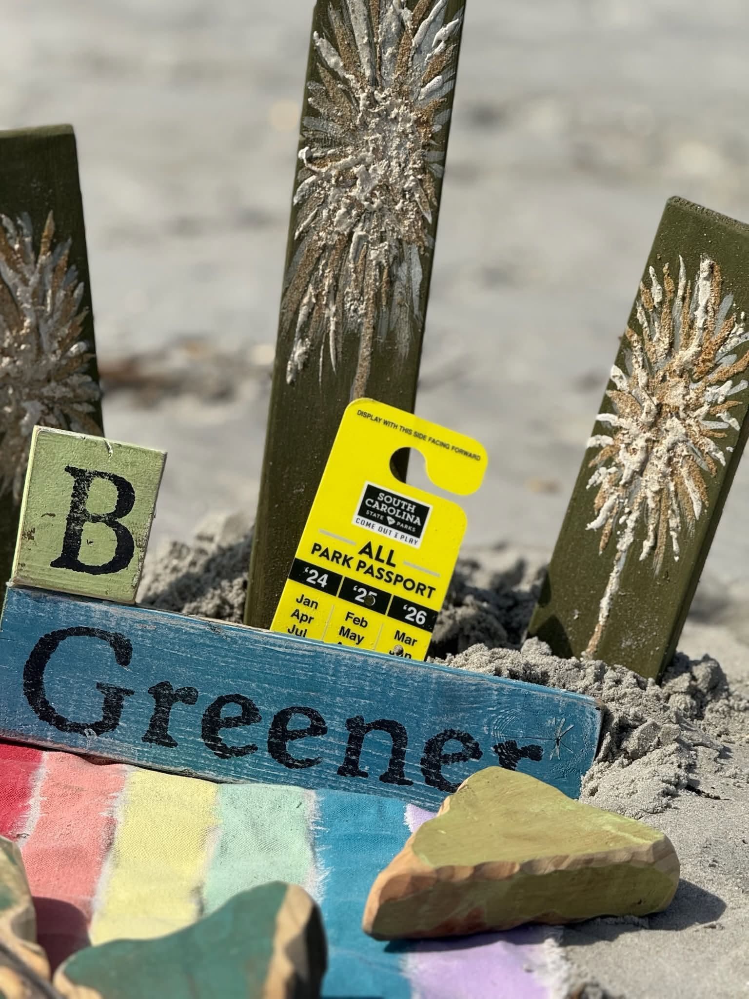 Colorful painted stakes with letters and a yellow tag in sand, one stone resting on a blue sign reading “Greene”