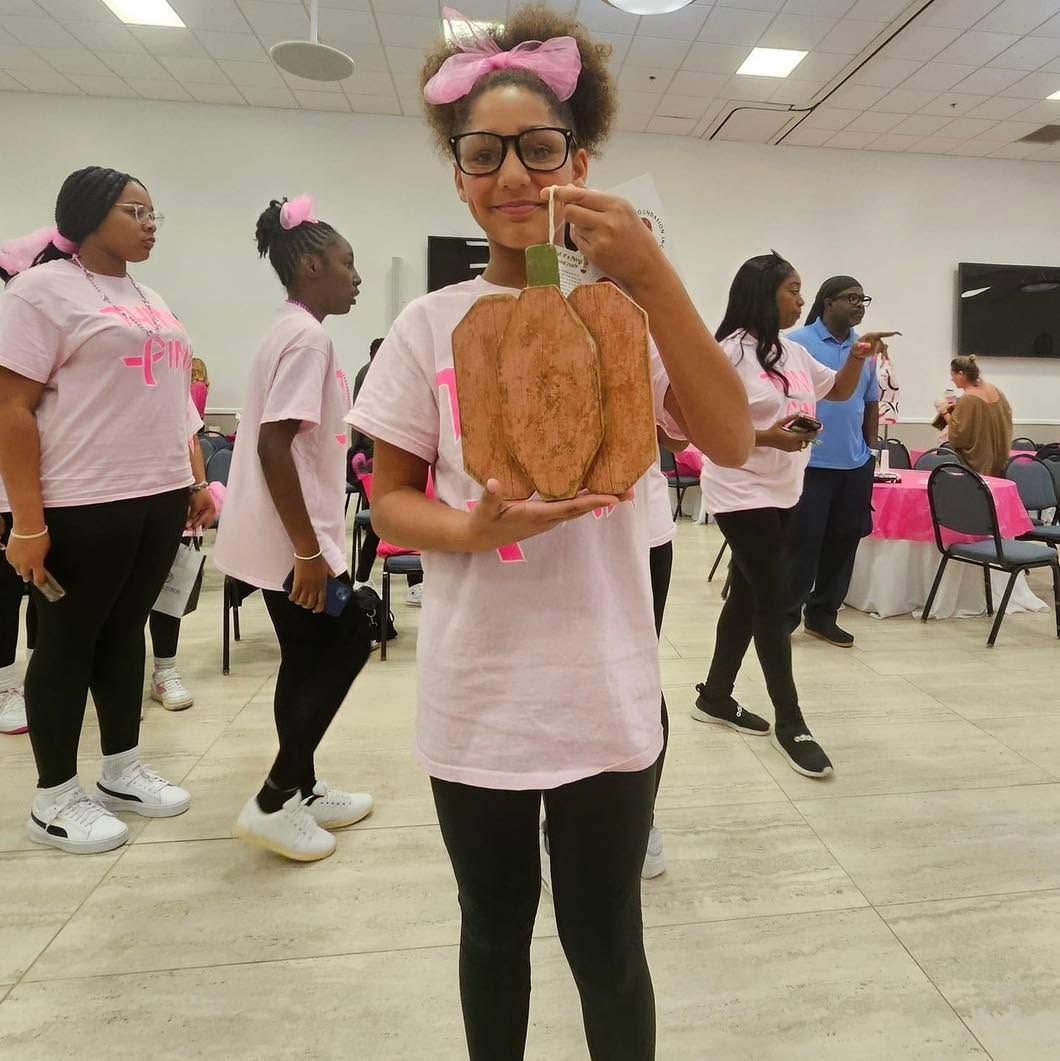 Girl in pink shirt holding a paper craft bag at an indoor event hall, with others in matching shirts behind her