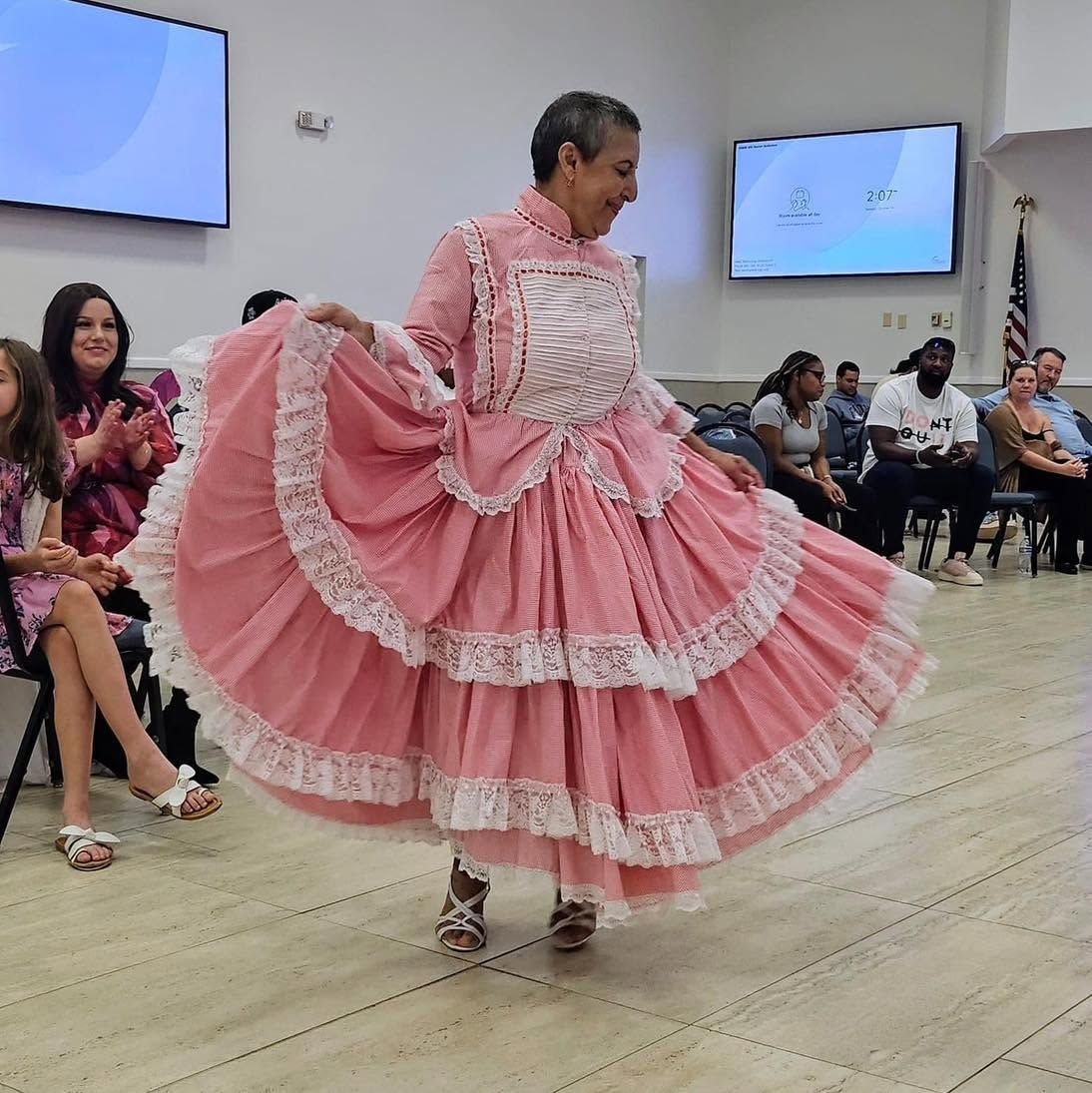 Woman in a pink ruffled dress dancing at an indoor event with seated audience and wall screens.