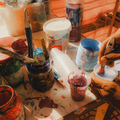 Hands mixing paint in a cluttered art workspace with colorful containers and splatters.