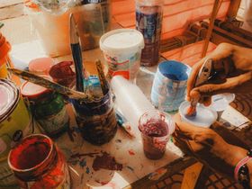 Hands painting colorful mugs and cups on a messy table with brushes, jars, and paint stains.