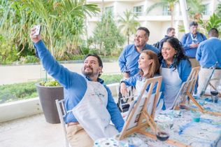 Group of people painting outdoors, smiling and taking a selfie at an easel in a bright courtyard.