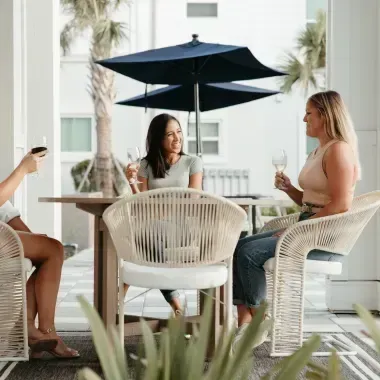 Three women at a table outdoors, holding wine glasses, under a blue umbrella.