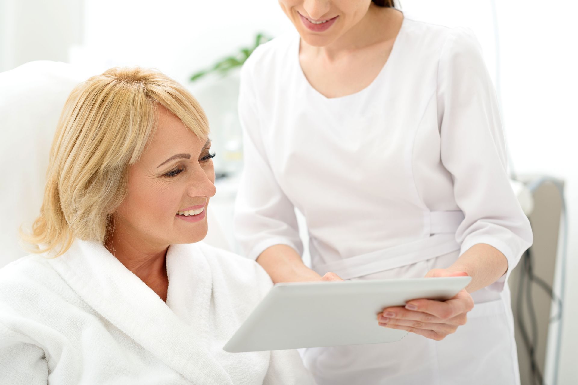 Dans un décor lumineux, un professionnel en uniforme blanc présente une tablette numérique à une personne souriante vêtue d'une robe blanche.