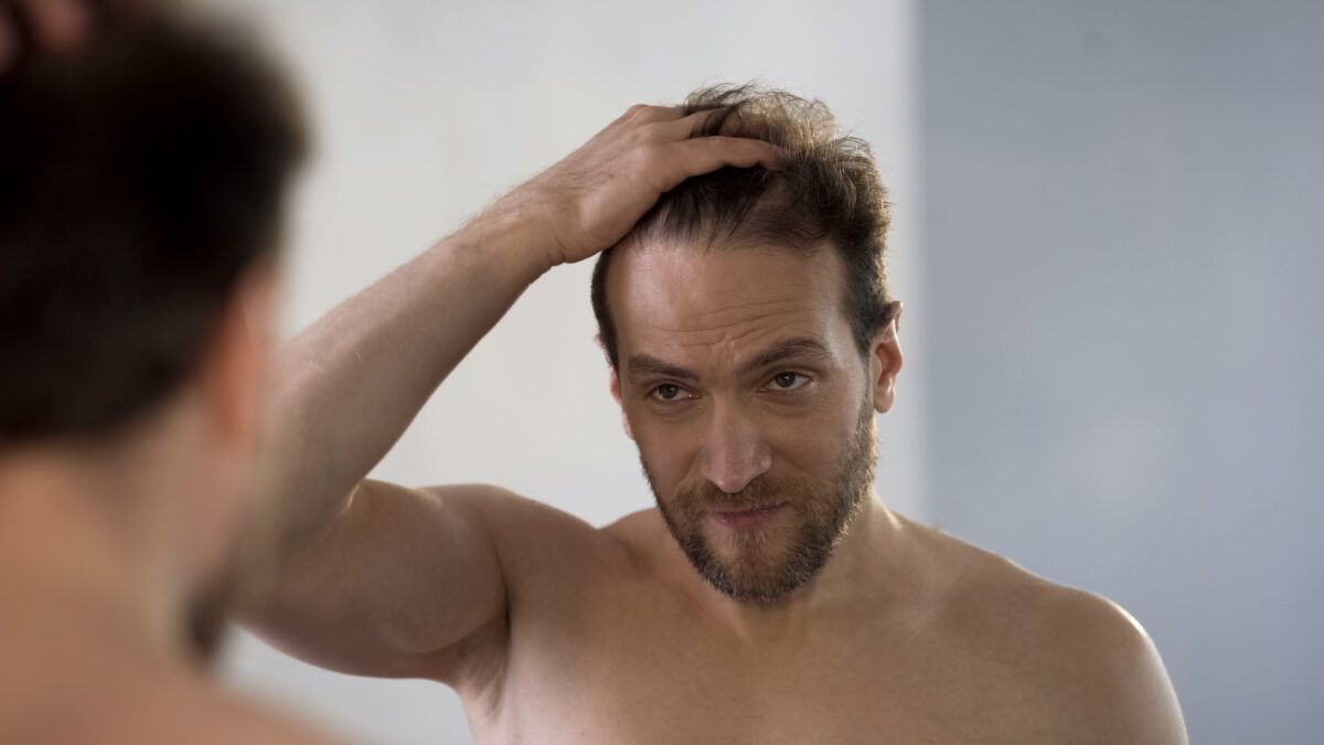 Une personne barbue se tient devant un miroir, touchant ses cheveux tout en regardant son reflet.