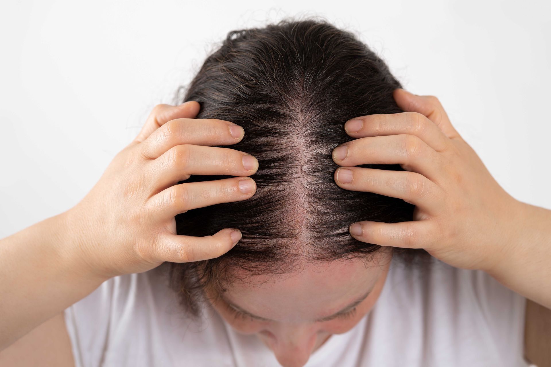 Person with dark hair examining scalp, likely checking for hair loss. Hands on head, white background.