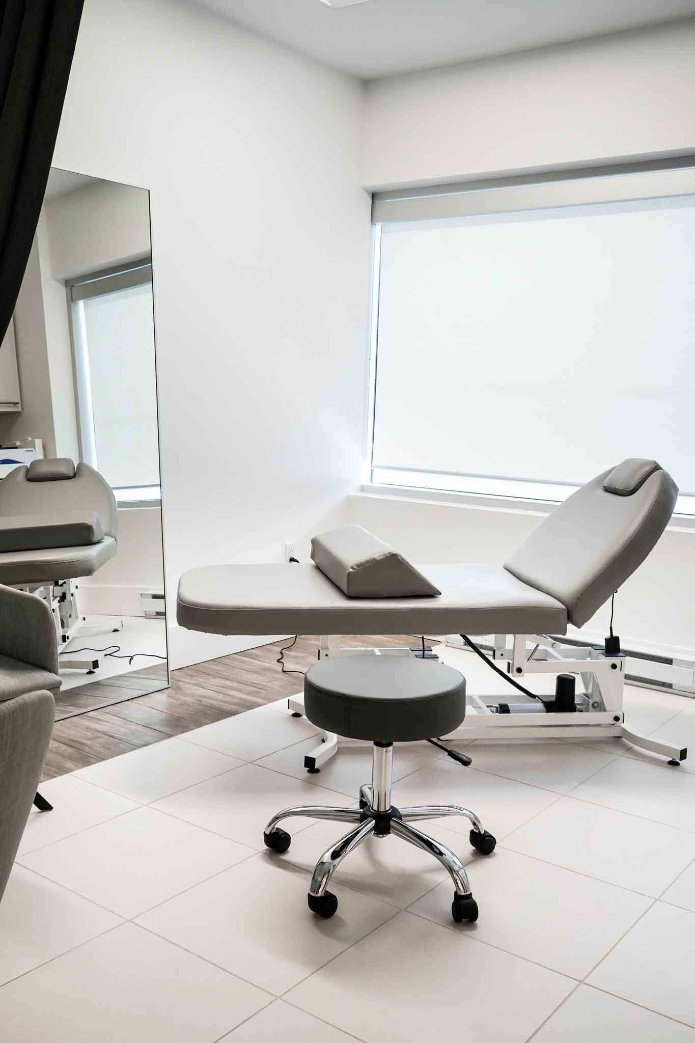 A treatment room featuring a light gray adjustable table, a rolling stool, and a tall mirror against a white wall.