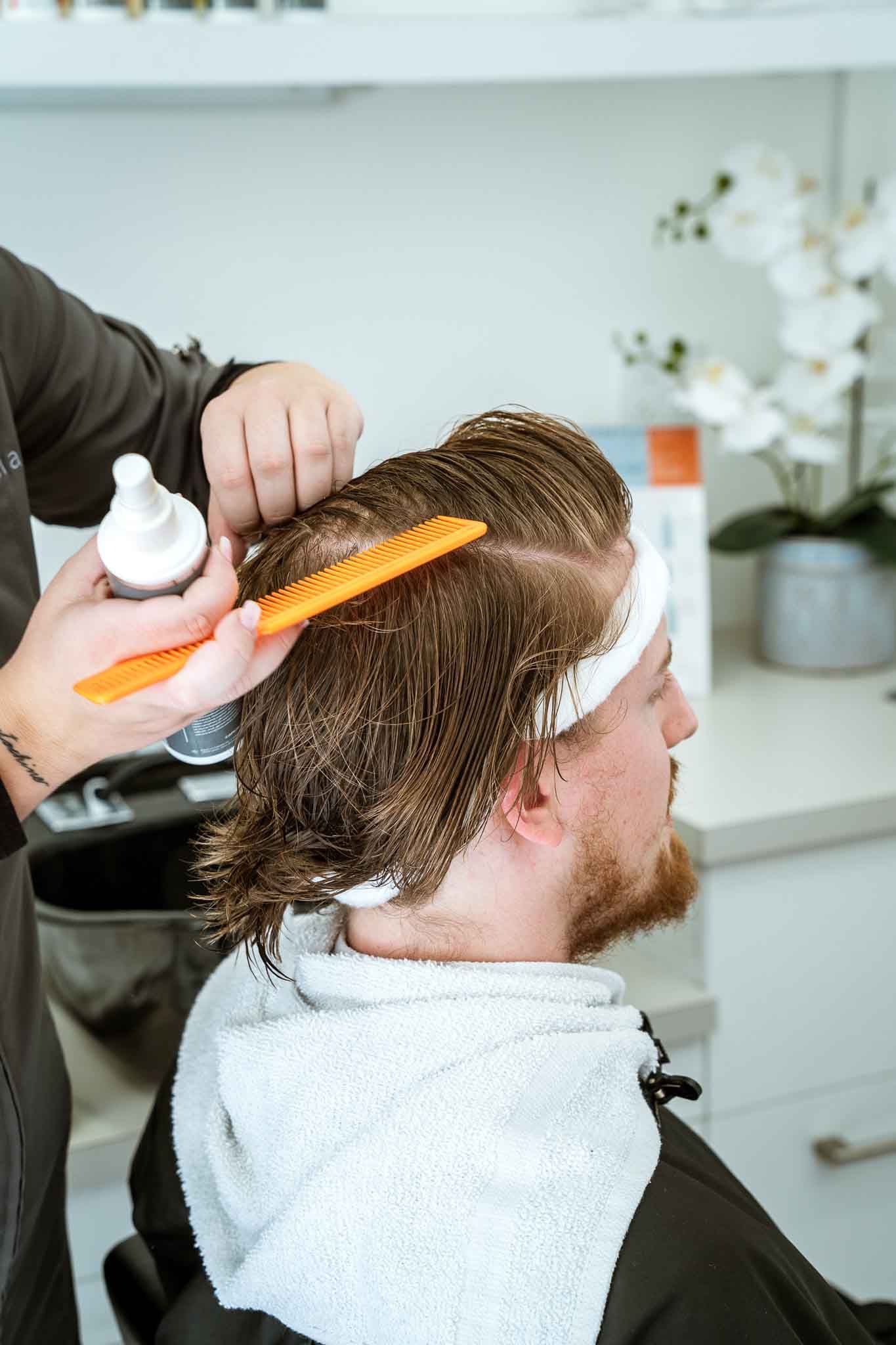 A stylist applies hair product to a person's damp hair with a bright orange comb in a salon setting.
