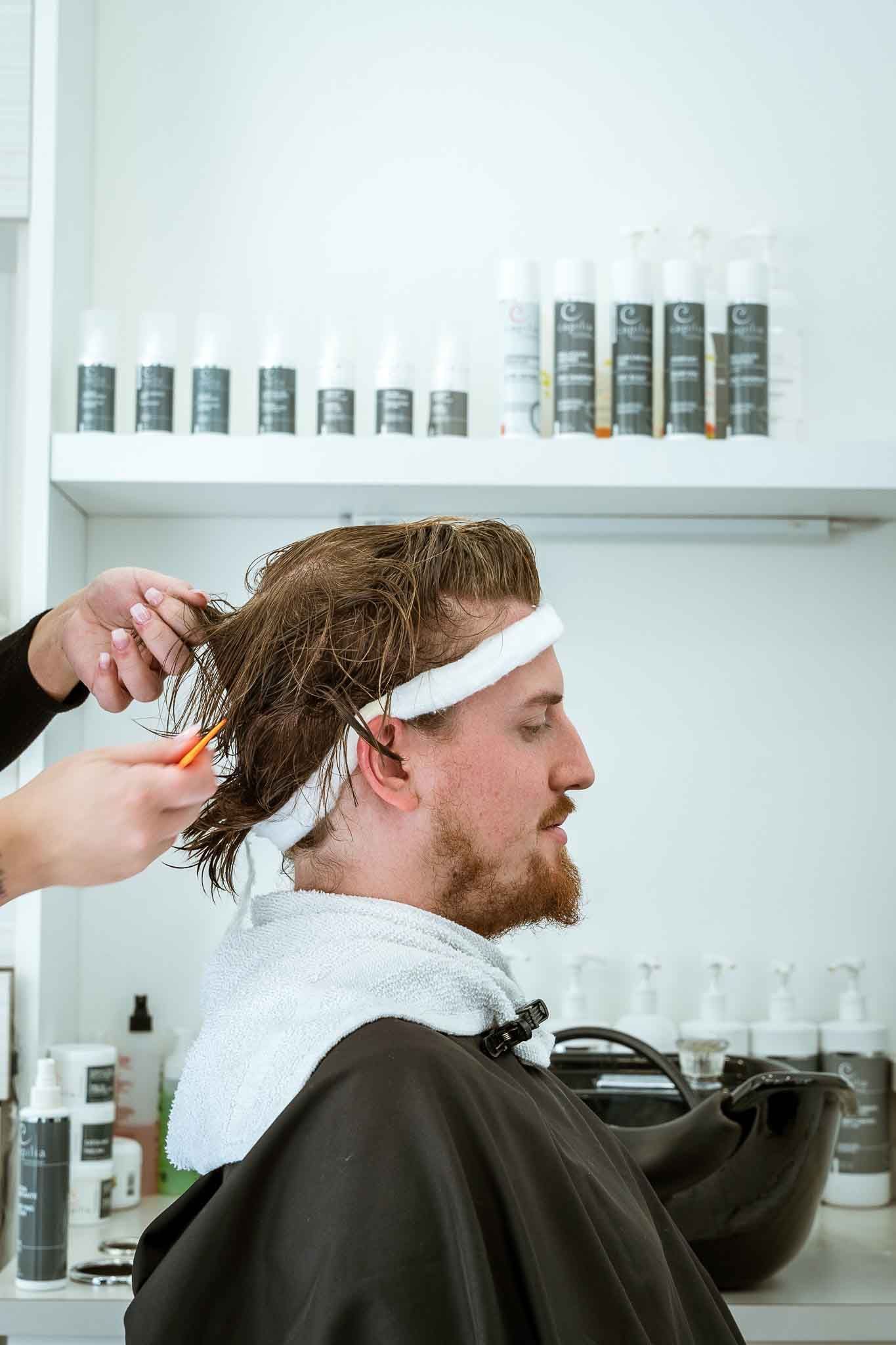 Dans un salon de coiffure, un coiffeur coupe les cheveux bouclés d'une personne, avec des étagères de produits capillaires professionnels en arrière-plan.