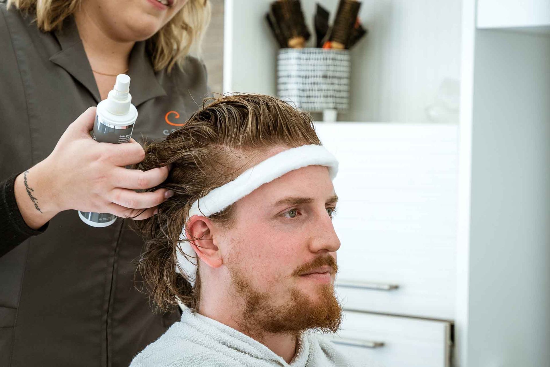 A stylist sprays a client's damp hair in a salon, while the client wears a white headband and cape.