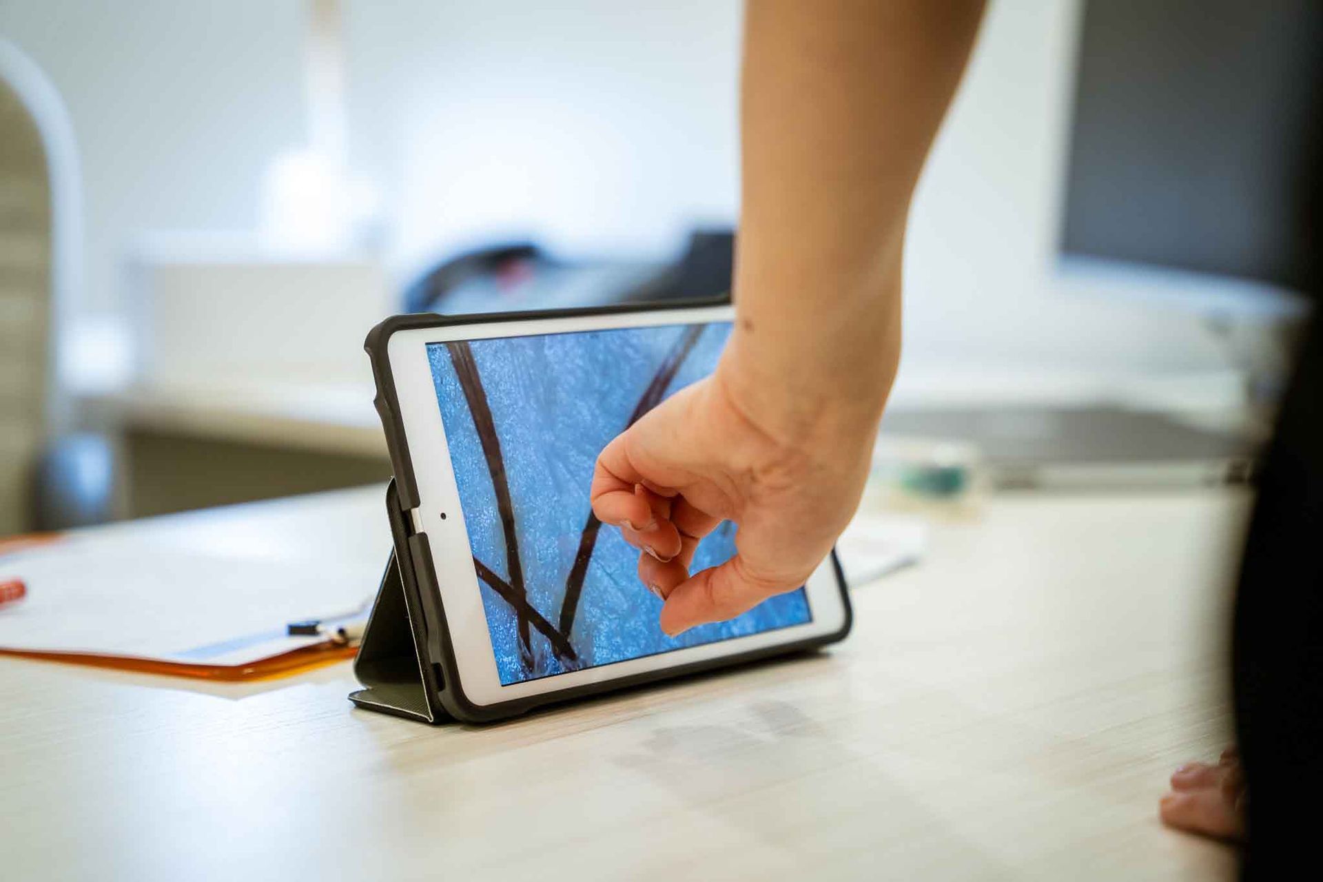 Une main désigne l'écran d'une tablette numérique affichant un motif abstrait de lignes sombres sur un fond bleu texturé.