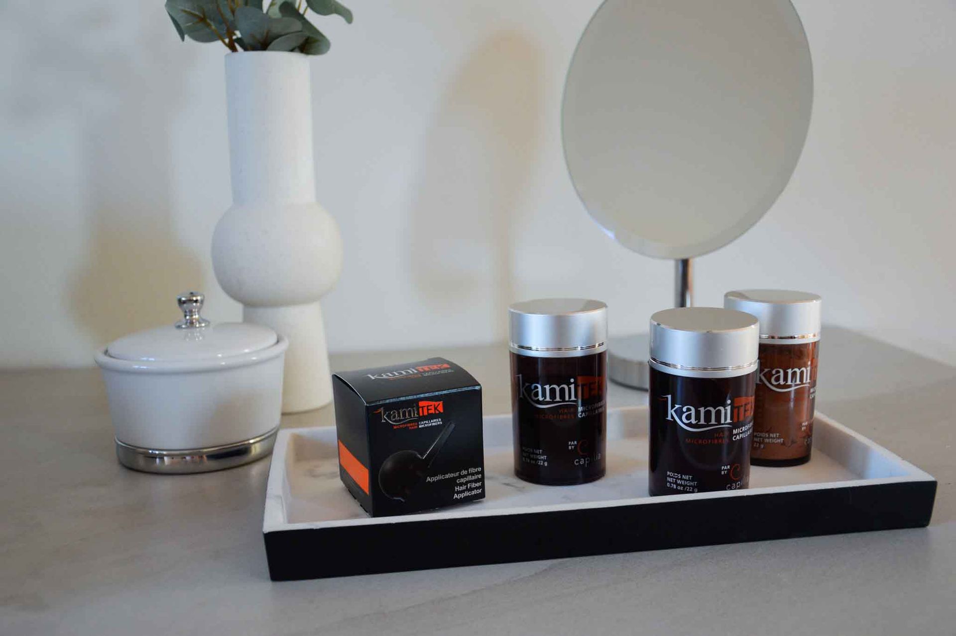 Sur un comptoir, un plateau blanc supporte des flacons de cosmétiques et une boîte, avec un miroir et un vase en arrière-plan.