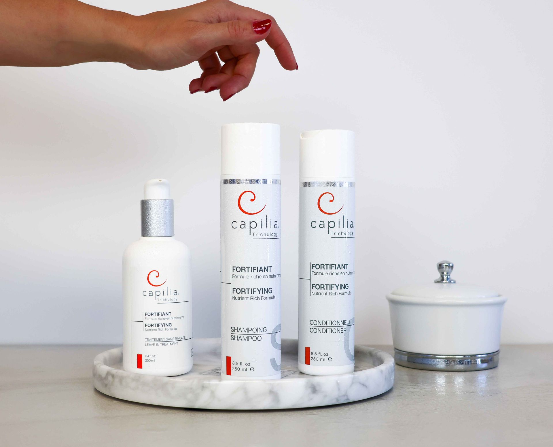A person's hand reaches toward three white bottles with red logos on a marble tray, next to a small white jar.
