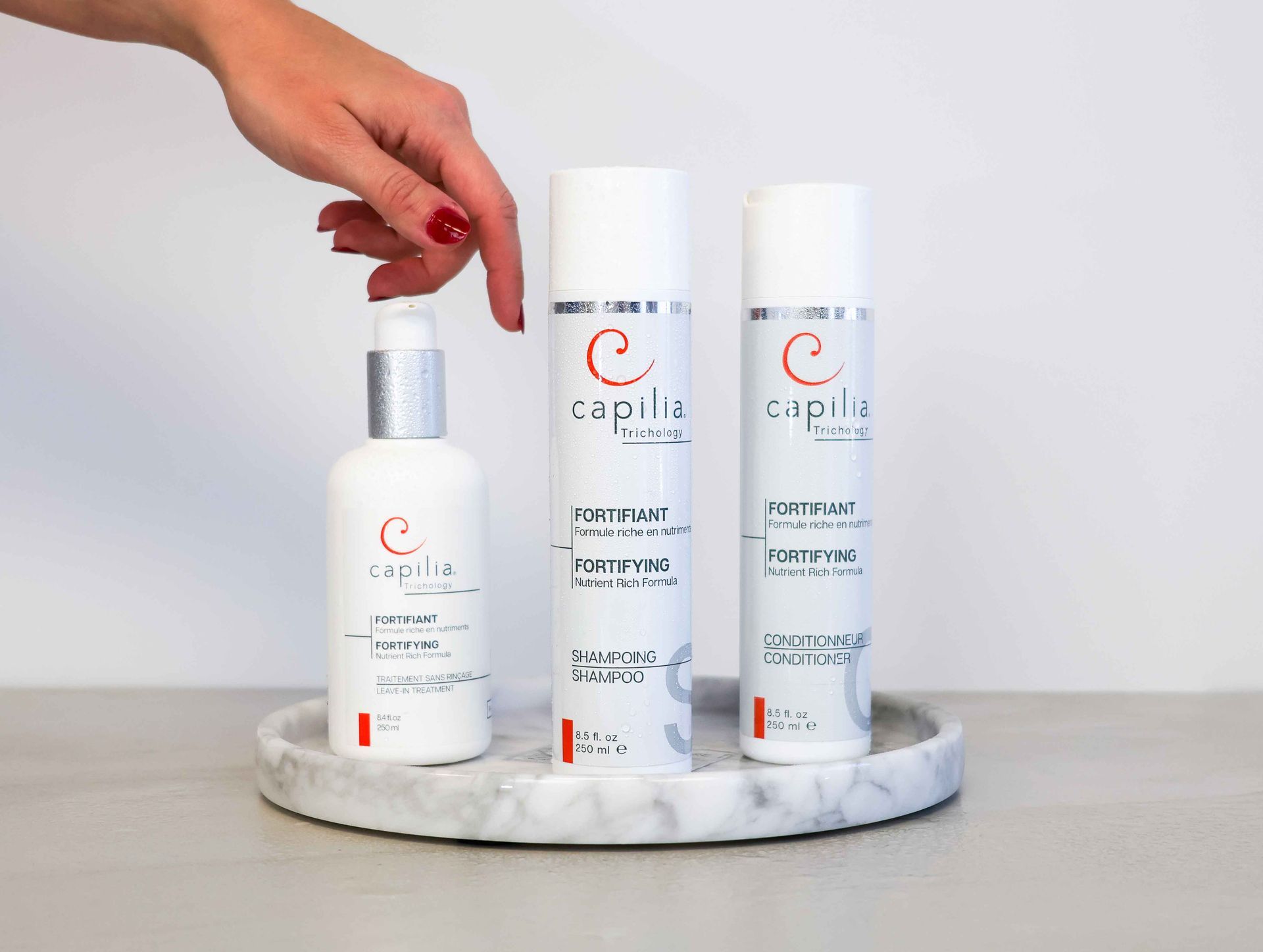 A hand places a white bottle next to two similar hair product containers on a small marble tray against a white wall.