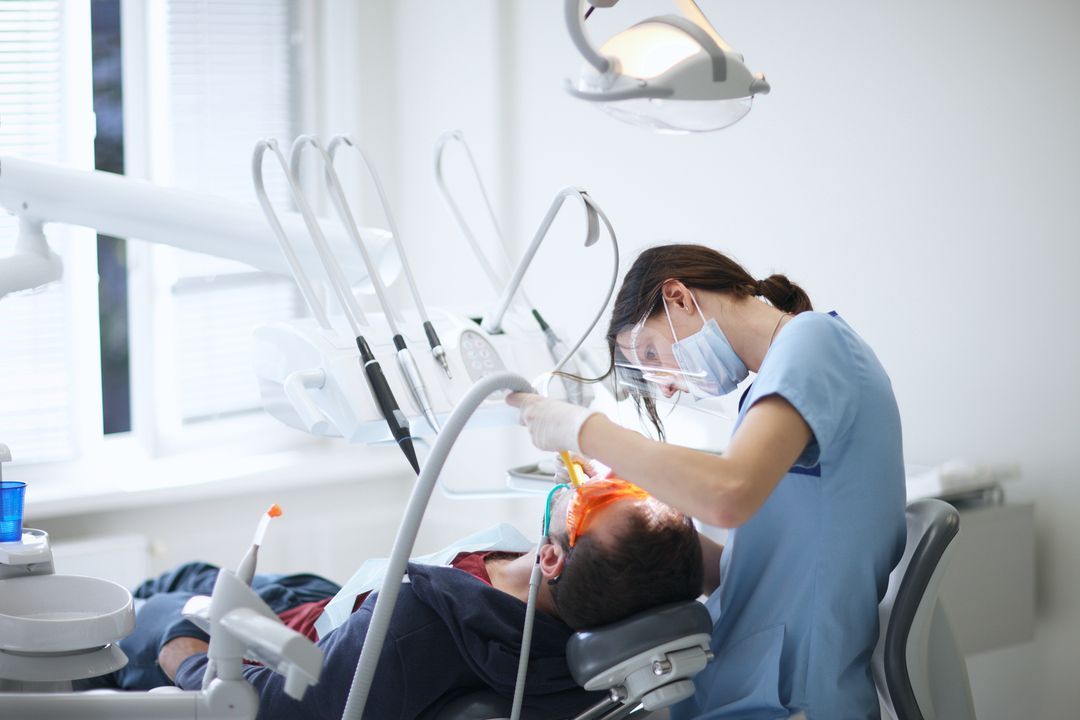 Dentist treating a patient in a bright dental clinic, leaning over the chair with tools and overhead lamp.