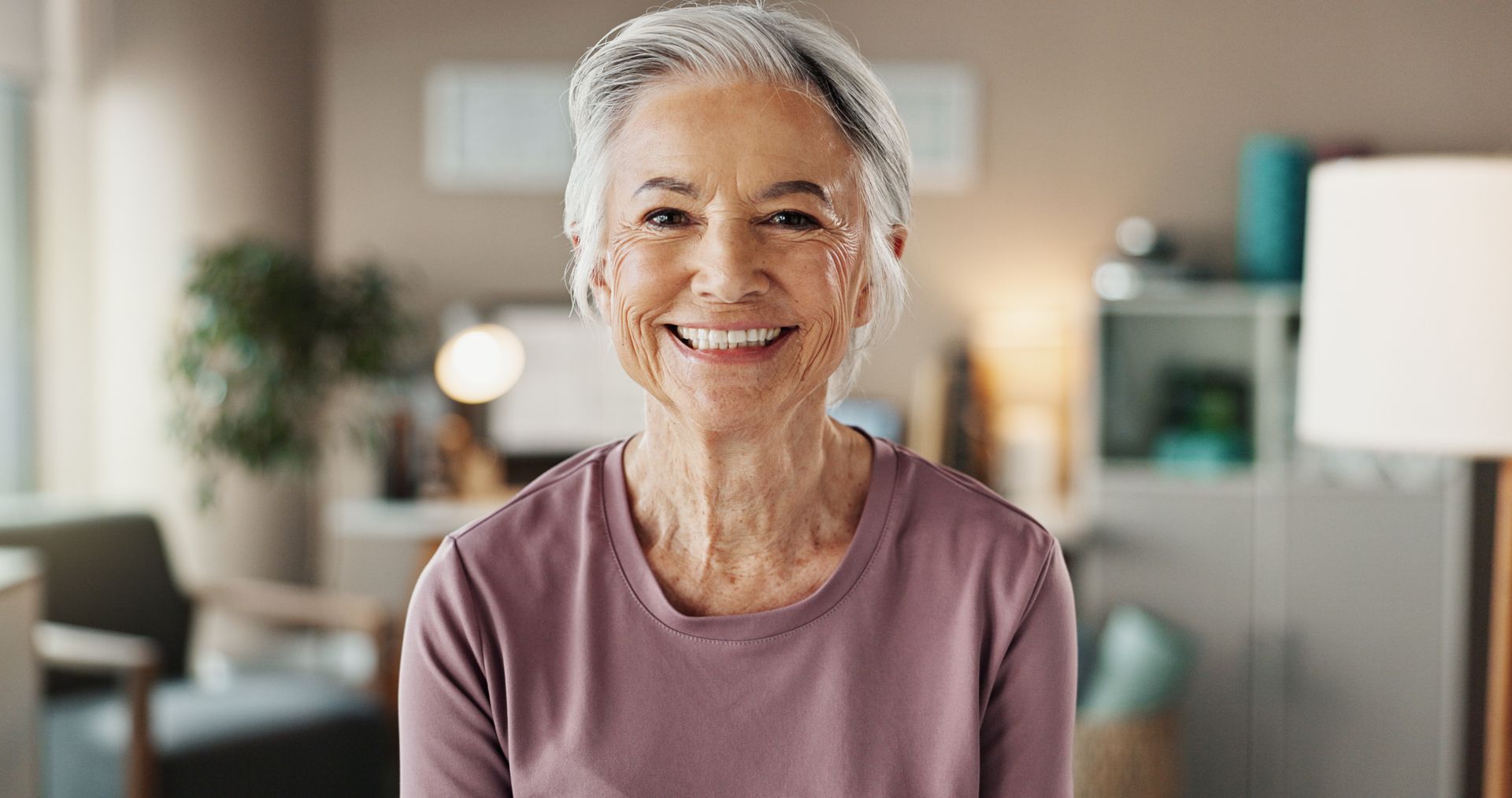 Smiling woman in a mauve top seated indoors with a softly blurred home background
