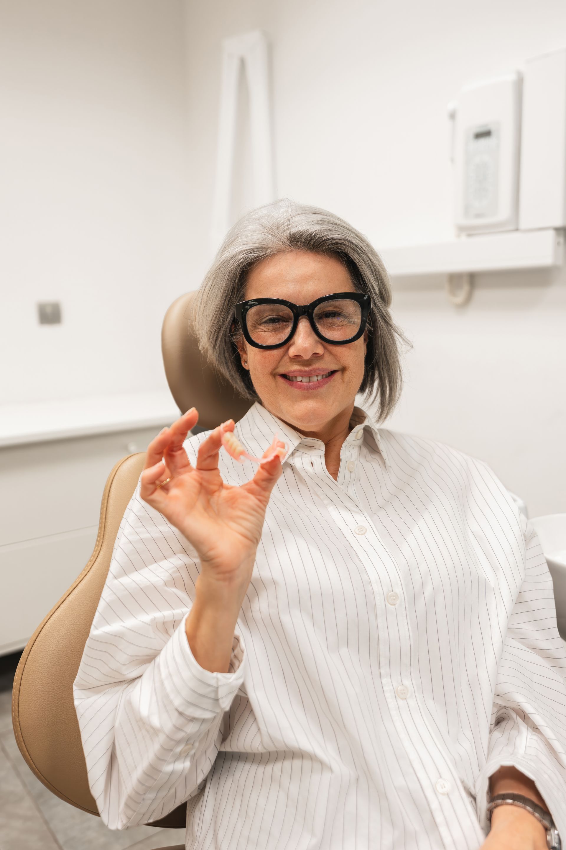 Person with gray hair and glasses seated in a clinic chair, gesturing with a small item in hand.