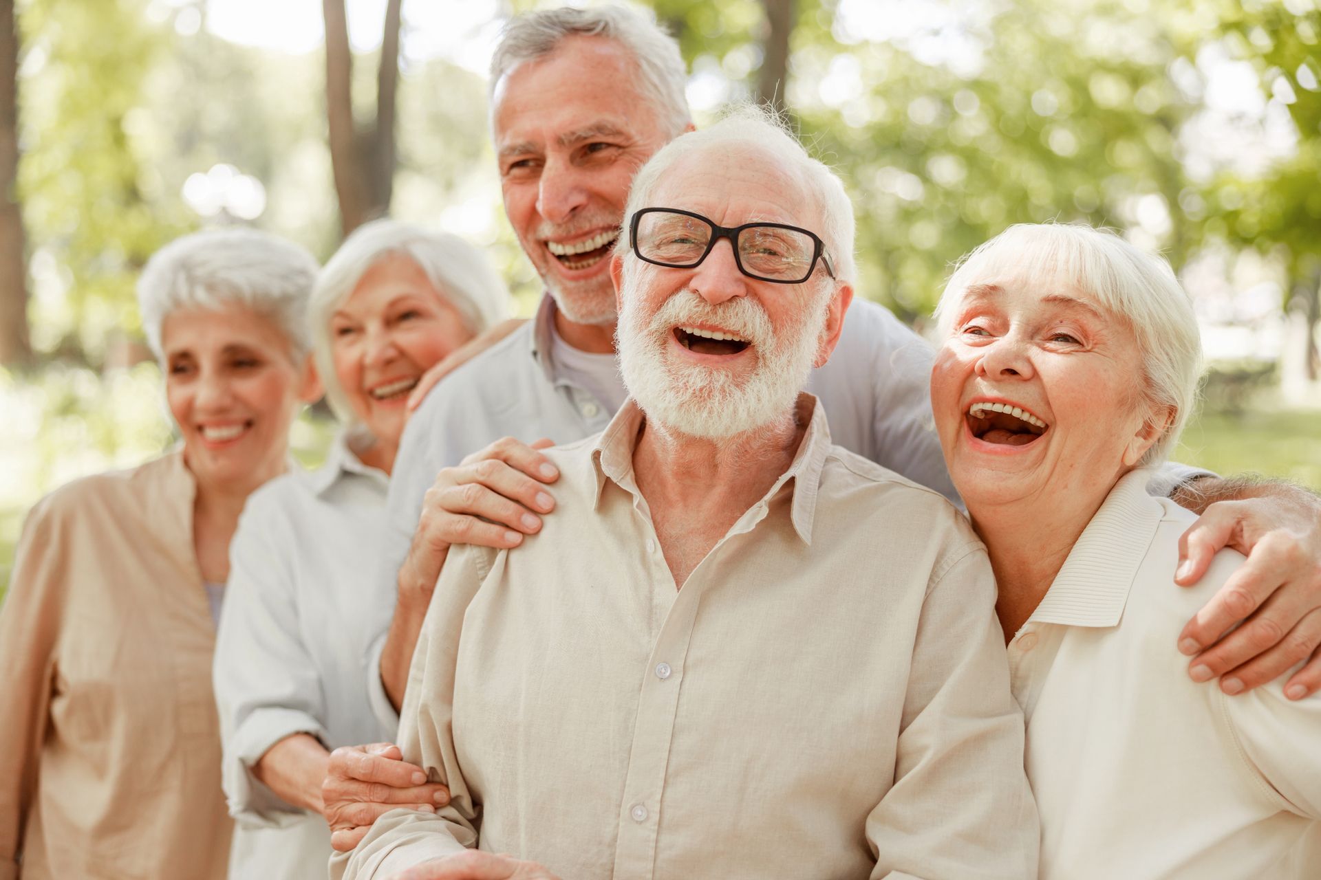 Group of smiling people outdoors, with an older man in glasses centered and others embracing behind him.