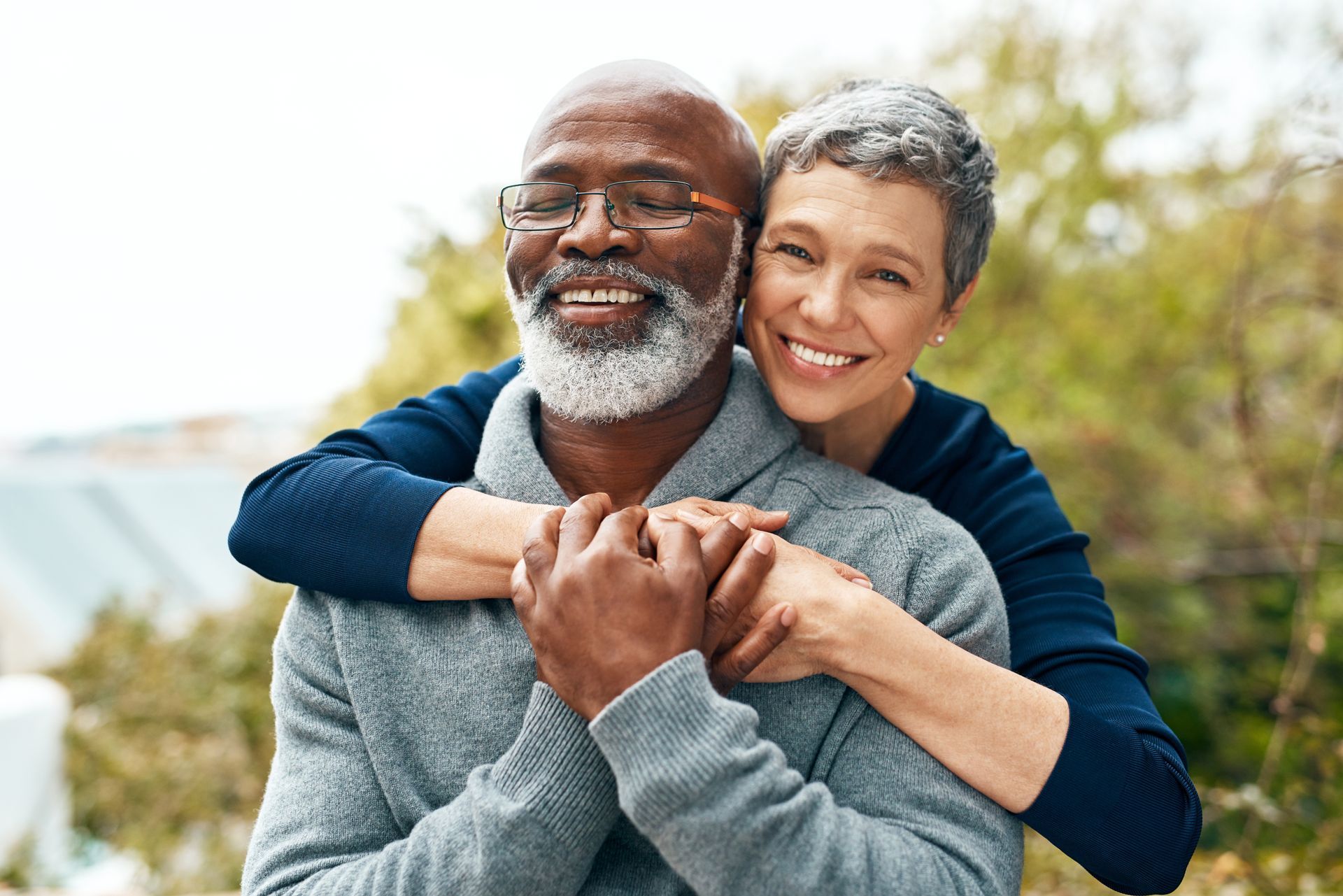 Smiling couple embracing outdoors in a grassy field, with a blurred coastline in the background.